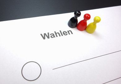 Drei Spielfiguren schwarz-rot-gelb auf einem Papier "Wahlen" stehend